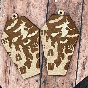 May include: A pair of wooden Halloween-themed earrings shaped like coffins. The coffins are decorated with a witch flying on a broomstick, bats, and a house.