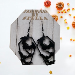 May include: Black acrylic earrings shaped like coffins with bats inside. The earrings are hanging from a white wooden display with the text "Treasures by Stella".