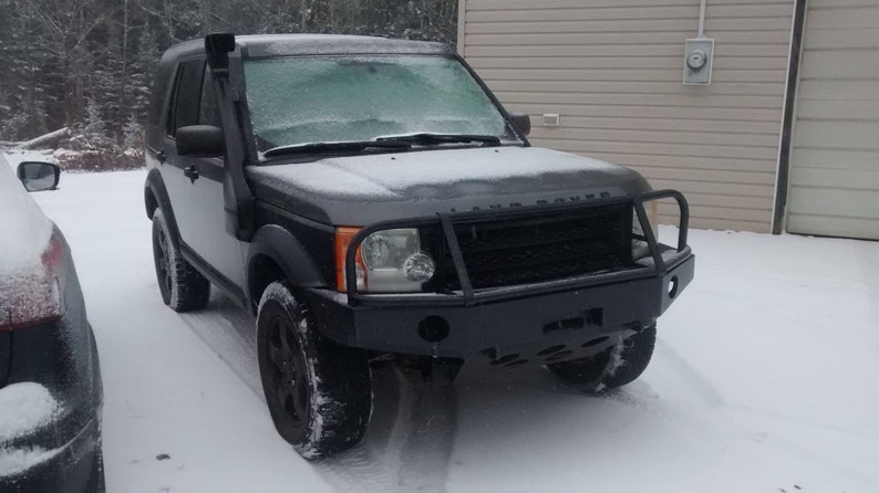 May include: A black Land Rover SUV with a front bumper guard and a snorkel, parked in the snow. The vehicle has black wheels and a black grille. The windshield is covered in frost.
