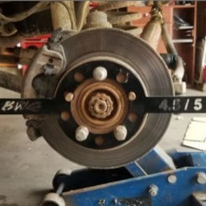 May include: A black metal tool with the text "BWKZ" and "4.5/5" printed on it. The tool is used to measure the lug pattern of a car wheel. The tool is placed on a car wheel with a black brake rotor and five lug nuts.