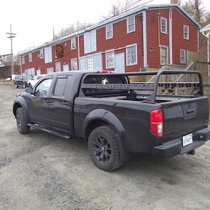 May include: Black pickup truck with a black truck bed rack. The truck is parked in front of a red brick building.