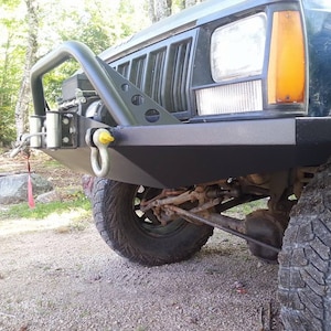 Op de afbeelding: Een zwarte Jeep met een aangepaste voorbumper en een lier. De bumper heeft een D-ring en een gele veiligheidspen. De Jeep staat geparkeerd op een grindpad.