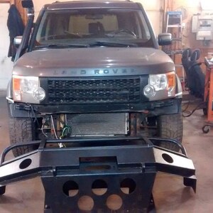 May include: A gray Land Rover SUV with its front bumper removed, revealing the radiator and engine components. A black metal bumper is positioned in front of the vehicle. The scene is set in a garage or workshop.