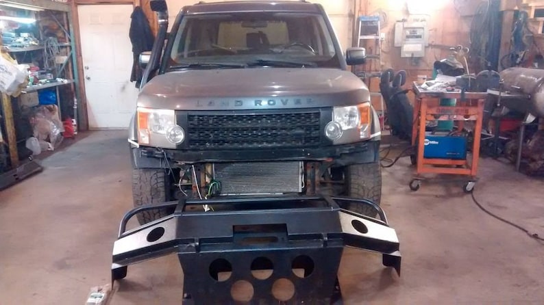 May include: A gray Land Rover SUV with its front bumper removed, revealing the radiator and engine components. A black metal bumper is positioned in front of the vehicle. The scene is set in a garage or workshop.