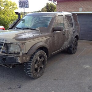 May include: A mud-covered Land Rover Discovery SUV parked on a paved driveway. The vehicle has a black front bumper, off-road tires, and a snorkel. The car is covered in mud, suggesting off-road use.
