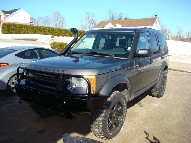 May include: A dark gray Land Rover SUV with a black front bumper guard and off-road tires. The vehicle has a snorkel and is parked outside on a sunny day. A silver car is parked next to it.