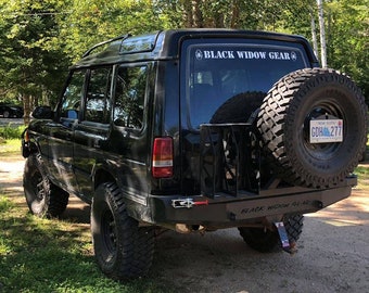 May include: Black Land Rover Discovery with a spare tire mounted on the back. The vehicle has a black bumper with a "Black Widow Gear" sticker on it. The license plate reads "GDH 277".