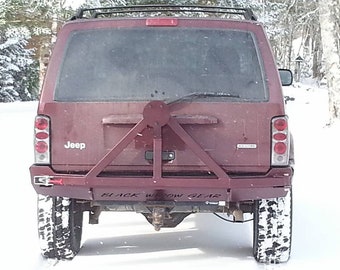 Può includere: Una Jeep Cherokee rossa con un paraurti in metallo nero e un portapacchi per pneumatici. Il portapacchi è un telaio triangolare con un pneumatico di scorta montato su di esso. Il paraurti ha la scritta "BLACK WIDOW GEAR" stampata su di esso.