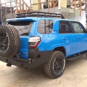 Könnte beinhalten: Ein blauer Toyota 4Runner Trail Edition mit einem Dachgepäckträger, einem Reserverad am Heck und schwarzen Felgen mit General Grabber Reifen.