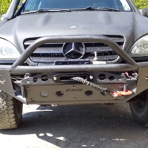 May include: Black off-road vehicle with a custom front bumper, winch, and brush guard. The bumper has a matte finish and integrated lights. The tyres are large and rugged, suitable for off-road driving.