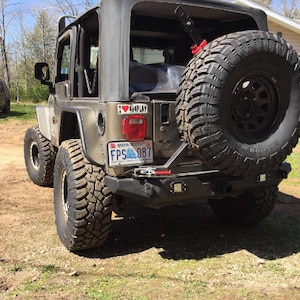 May include: A silver Jeep Wrangler with a spare tyre mounted on the back. The Jeep has a black bumper with a tow bar and a number plate that reads "FPS 087". The Jeep also has a sticker that says "I love 4x4 play".