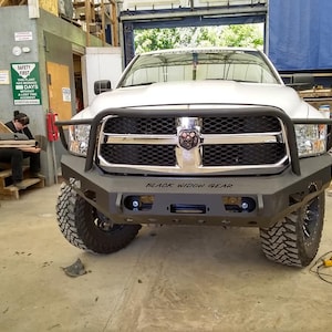 Peut inclure: Un camion pick-up argenté avec un pare-chocs avant noir et une calandre noire. Le pare-chocs est doté d'une barre de protection en acier noir avec le logo "Black Widow Gear". Le camion est garé dans un garage.