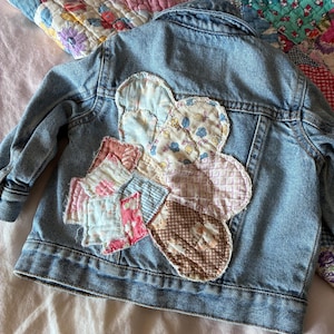 May include: A light blue denim jacket featuring a large, multi-colored floral-shaped patch on the back. The patch is constructed from various quilted fabric squares in shades of pink, blue, brown, and white, showcasing diverse patterns.