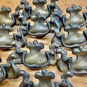May include: A collection of antique brass drawer pulls. Each pull features a curved handle and decorative top, with a tarnished finish. The hardware is arranged on a wooden surface, showcasing their vintage aesthetic.