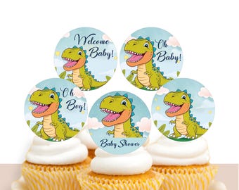 Könnte beinhalten: Cupcakes mit weißem Zuckerguss und gelben Papierförmchen, gekrönt von runden Dinosaurier-Dekorationen. Die Dekorationen zeigen einen grünen Dinosaurier und die Aufschriften "Welcome Baby!", "Oh Baby!", "Oh Boy!" und "Baby Shower".