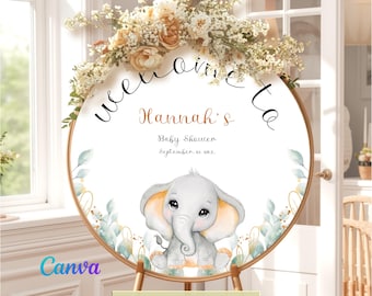 May include: A circular welcome sign for a baby shower featuring a watercolor illustration of a gray elephant with large ears. The text reads Welcome to Hannah's Baby Shower September 25, 20xx. The sign is decorated with floral arrangements.