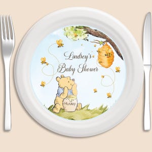 May include: White paper plate with a Winnie the Pooh baby shower design. The plate features Winnie the Pooh holding a jar of honey, a beehive, and bees flying around. The text on the plate reads "Lindsey's Baby Shower".