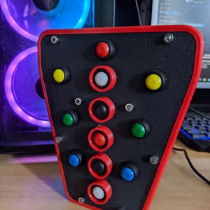 May include: A black and red gaming controller with multiple buttons, including red, yellow, green, blue, and white buttons. The controller is mounted on a black base.