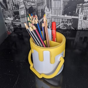 May include: A yellow paint can shaped pencil holder with a white interior. The holder has a black and white striped handle and is filled with coloured pencils and pens.