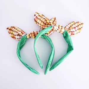 Può includere: Due fasce per capelli verdi con fiocchi in tessuto a forma di pannocchia di mais. I fiocchi sono annodati e le fasce sono realizzate in un materiale morbido ed elastico.