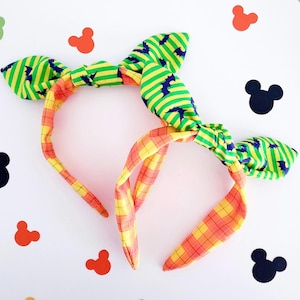 May include: Two colourful headbands with knotted bows. One headband is yellow and orange plaid, the other is green and yellow with a black bat pattern.