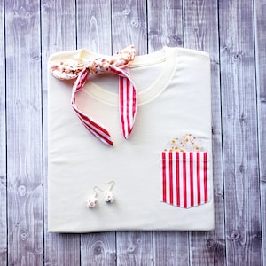 Puede incluir: Una camiseta blanca con un bolsillo a rayas rojas y blancas. El bolsillo tiene un diseño floral. La camiseta también tiene una pajarita con estampado rojo y blanco.