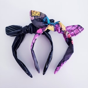 May include: A black and white striped headband with a colourful patterned bow. The bow features a patchwork design with pink, yellow, and purple accents.