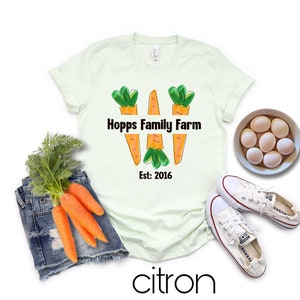 May include: A light green t-shirt featuring a graphic of three orange carrots with green tops. The text "Hopps Family Farm" is printed in black above the carrots and "Est: 2016" is printed in black below the carrots.