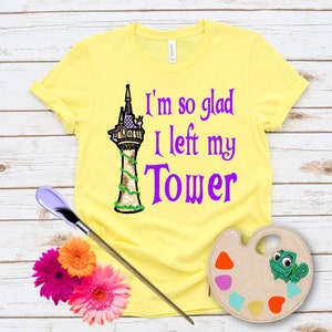 May include: A yellow t-shirt with the text "I'm so glad I left my Tower" in purple. The shirt features a cartoon illustration of a tower with green vines. A paintbrush, flowers, and a palette are also in the image.