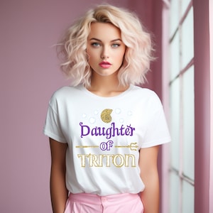 May include: White t-shirt with gold glitter text that reads "Daughter of Triton" with a gold seashell and a trident graphic.