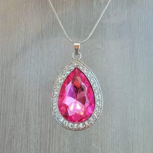 May include: A silver necklace featuring a large, teardrop-shaped pendant. The pendant has a vibrant pink gemstone at its center, surrounded by a halo of small, clear crystals. The chain is delicate and the pendant is a statement piece.