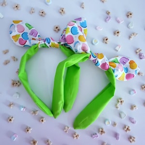 May include: Two playful headbands featuring bunny ears. The ears are patterned with colourful polka dots, and the headband and long, floppy ears are vivid green. The headbands are surrounded by small, decorative sweets.