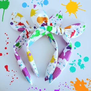 May include: Two white headbands with a colorful paint splatter design. The headbands have a bunny ear shape and are decorated with splashes of red, orange, yellow, green, purple, and blue paint. The background is white with additional paint splatters.
