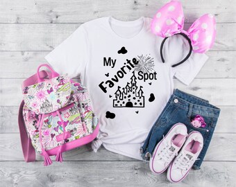 Puede incluir: Camiseta blanca con texto negro que dice "My Favorite Spot" y una silueta negra de un castillo con fuegos artificiales. La camiseta se combina con una diadema de lunares rosas, una mochila estampada en rosa y blanco, pantalones cortos de mezclilla azul y zapatillas blancas.