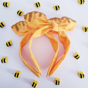 May include: A yellow and gold headband with a honeycomb pattern and a bow. The headband is decorated with a gold glitter finish.