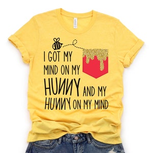Puede incluir: Camiseta amarilla con un bolsillo rojo y un gráfico de abeja. El texto en la camiseta dice "I got my mind on my honey and my honey on my mind".