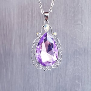 May include: A silver necklace with a large purple teardrop-shaped pendant. The pendant is surrounded by a silver filigree design and has smaller clear rhinestones.