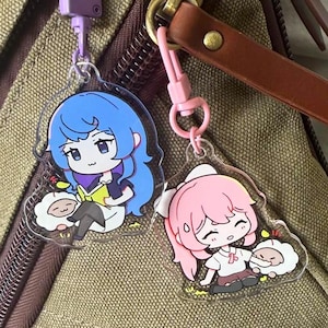 May include: Two acrylic charms attached to a brown canvas bag. One charm features a blue-haired character reading a book next to a small white creature. The other charm shows a pink-haired character sitting beside a similar white creature.