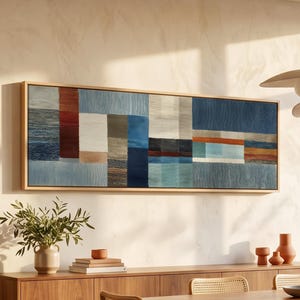 May include: A rectangular wall art piece featuring a horizontal abstract composition of woven-style blocks in shades of blue, white, gray, and rust orange, enclosed in a light-toned wooden frame, displayed above a wooden sideboard and dining table.