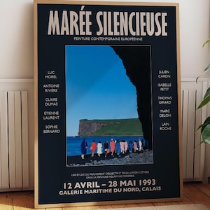 May include: A framed poster with the title "MARÉE SILENCIEUSE" in white text on a dark blue background. The poster features a coastal landscape with figures and text in French. The frame is a light brown color. The poster is leaning against a white wall.