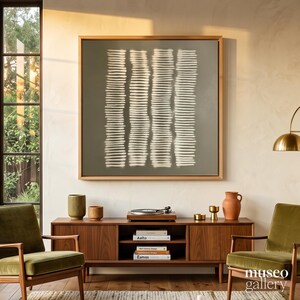 May include: A square framed wall art piece featuring four vertical columns of horizontal white brushstrokes on a muted olive green background, displayed above a wooden media console with books, ceramic vessels, and a record player.