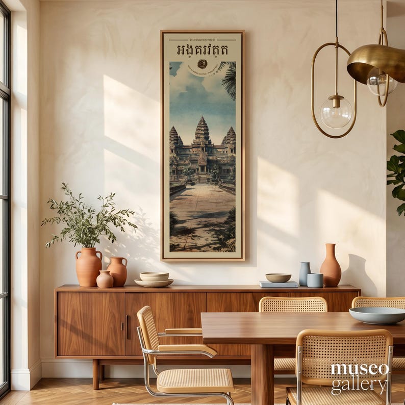 Pu&ograve; includere: Una stampa artistica da parete verticale incorniciata che raffigura il complesso del tempio di Angkor Wat in Cambogia. L'opera mostra la struttura in pietra sotto un cielo azzurro con nuvole, con testo khmer in alto. La stampa &egrave; in una cornice in legno chiaro.