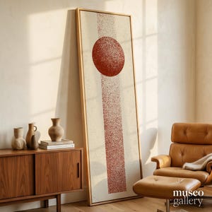May include: A tall, rectangular framed wall art piece featuring a textured, rust-colored vertical stripe and a solid circle centered on a cream-colored background. The artwork is displayed in a light wood frame, showcasing a grainy, stippled artistic effect.