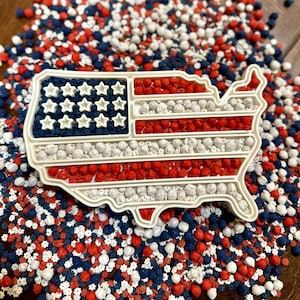 May include: A white plastic mold in the shape of the United States, filled with red, white, and blue candies, resembling the American flag. The mold is surrounded by a scattering of similar candies, creating a patriotic theme.