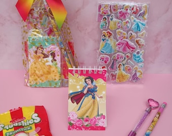 May include: A gift bag with a rainbow bow, notebooks, stickers, pencils, and candy. The notebooks and pencils feature Disney princess designs. The candy is a red and yellow package of Squishies Drumstick candy.