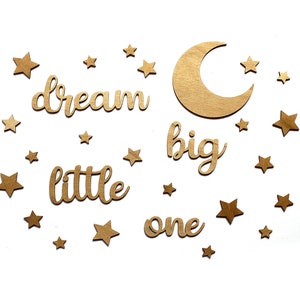 Può includere: Lettere in legno tagliate dorate che compongono la scritta "dream big little one" con una luna crescente e diverse stelle sparse intorno.