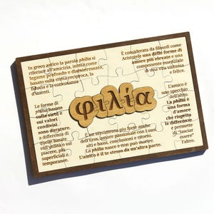 Può includere: Un puzzle in legno con una cornice marrone scuro. I pezzi del puzzle sono beige e presentano la parola greca "φιλία" in un carattere stilizzato. Il testo in italiano circonda la parola centrale, discutendo il concetto di philia, una forma di amore e amicizia.