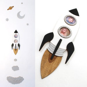 Pode incluir: Moldura de fotos de foguete branco com detalhes em preto e cinza. O foguete tem três ranhuras para fotos, duas redondas e uma retangular. O foguete tem uma base de madeira marrom. O texto "explorar" está escrito em uma nuvem abaixo do foguete.