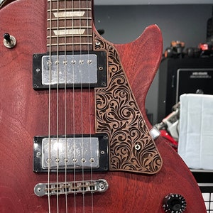 Puede incluir: Una guitarra eléctrica burdeos con un golpeador decorativo con un diseño floral grabado. La guitarra tiene perillas de control negras, pastillas plateadas y un diapasón de palisandro. Está montada en un soporte negro.
