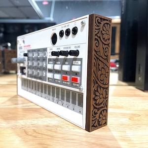 Può includere: Un Roland TR-8S Rhythm Composer bianco con un pannello laterale in legno che presenta un intricato design floreale. Il pannello è di colore marrone scuro con una venatura del legno marrone chiaro.